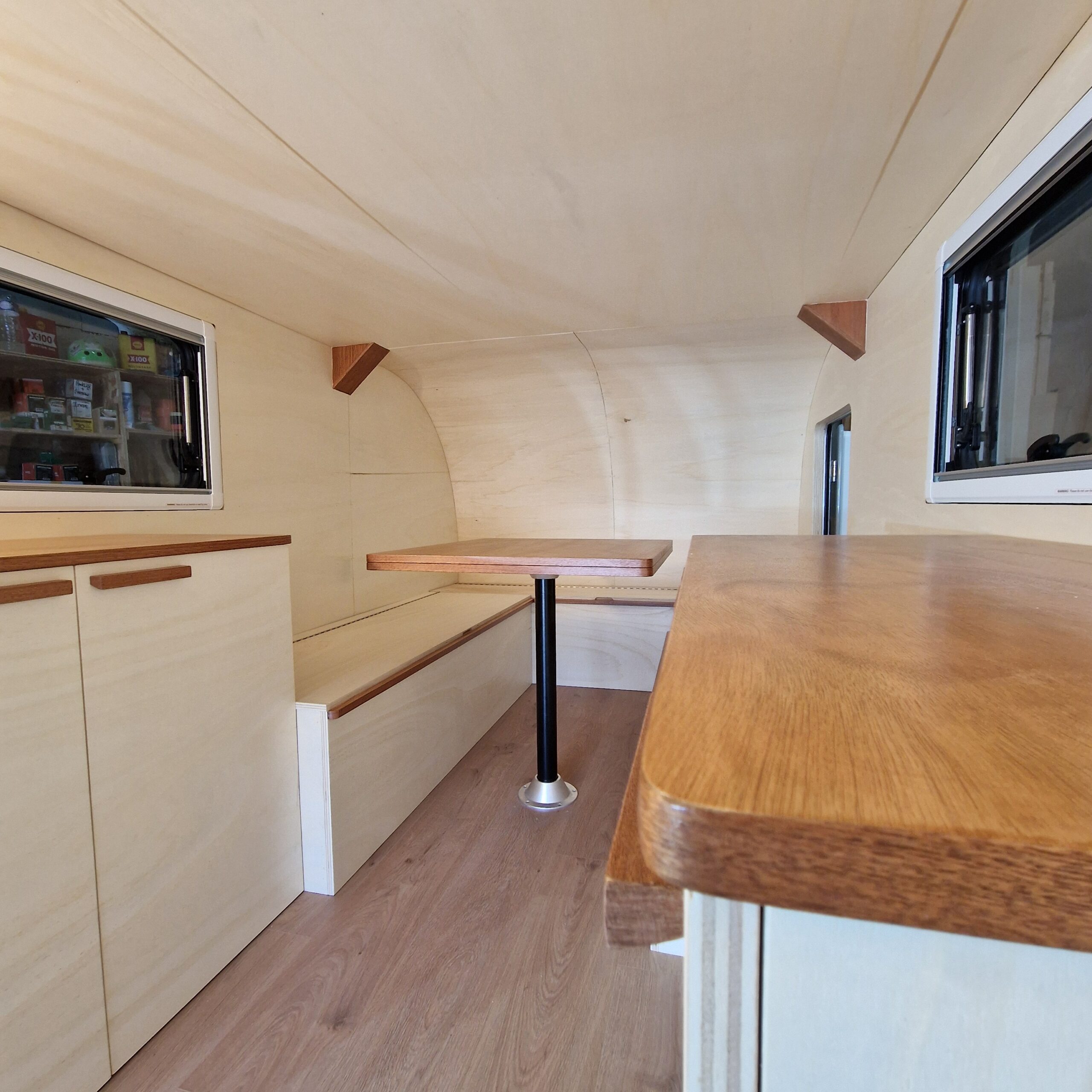 Aménagement de remorque à bordeaux avec banquette en U, table escamotable, meubles de rangement, habillage arrondie sur mesure et baies ouvrantes